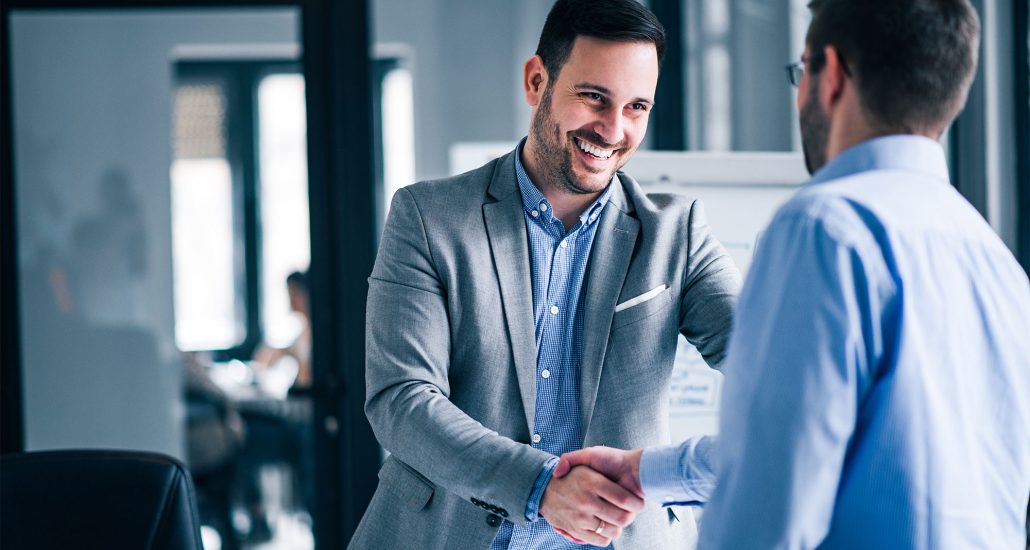 Two smiling businessmen shaking hands while standing in an office.